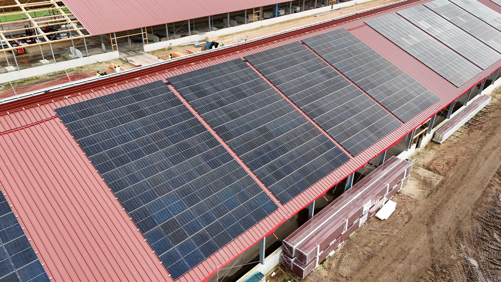 Nahaufnahme der ersten fertiggestellten Modulsektion auf dem roten Trapezdach, rechts Rohbaugerüst der zweiten Halle